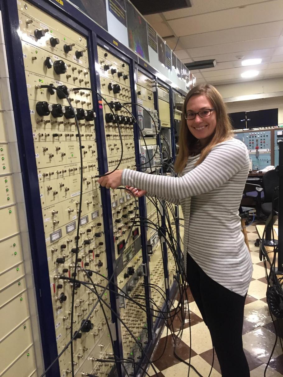 Woman stands in front of a large control board with multiple wires and access points. 