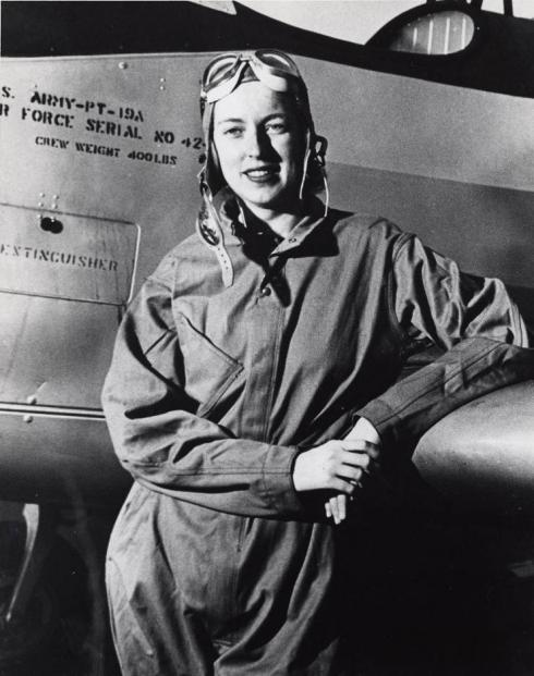 A woman in aviator gear leans against an airplane wing. 