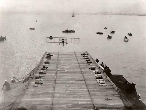 A Curtiss Model D flying toward a ship which has a makeshift runway. Other boats dot the water.