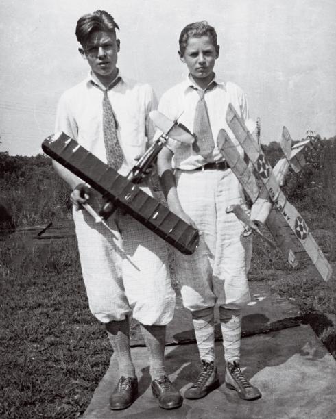 Two teenage boys, slightly rumpled but still wearing ties, stand outdoors, sometime in the 1930s, holding two rubber-band powered airplane models that they built.