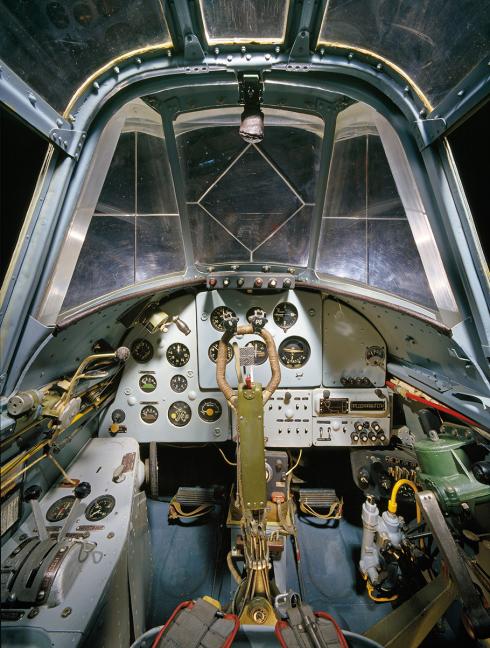 A view inside the cramped cockpit of the Museum’s Il-2 Shturmovik. A pilot’s seat is surrounded by multiple dials and exposed equipment, while facing a clear glass canopy.