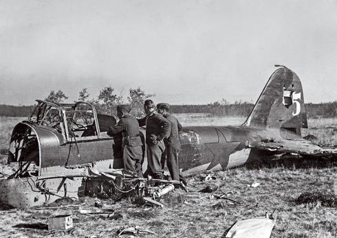 Three German soldiers inspect a shot-down IL-2, which sits in a field amid debris, missing its front and wings.