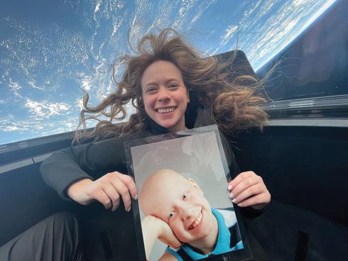 A young woman is in orbit aboard a Dragon space capsule. The Earth looms in the background while she holds up a photo of herself when she was a pediatric cancer patient.