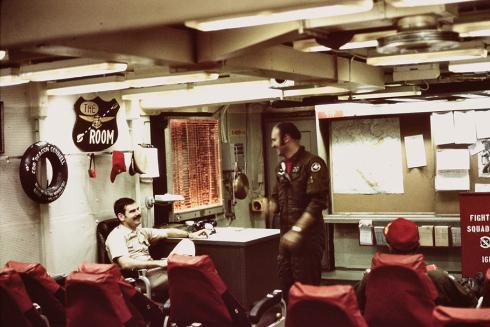 The ready room aboard Midway is cramped and illuminated by overhead fluorescent lights. Lieutenant Mike “Taco” Bell shares a laugh with Lieutenant Commander “Mugs” McKeown.