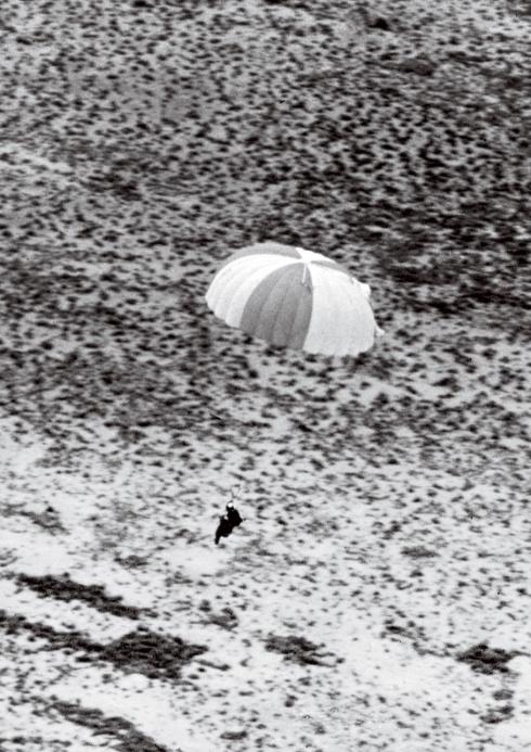  A grainy black and white photograph shows a figure using a deployed parachute above a landscape. 