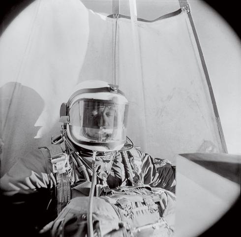 A black and white photograph Joseph M. Kittinger Jr. taken inside the gondola of Kittinger’s balloon. The sides of the balloon are visible behind him. He wears a suit and helmet akin to a spacesuit or high-altitude pilot’s suit for protection.