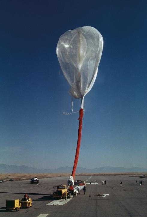 A semi-inflated silvery-white balloon rises in the air, attached to the ground by a large read tether. This is the beginning of the ascent in the Excelsior III gondola. 