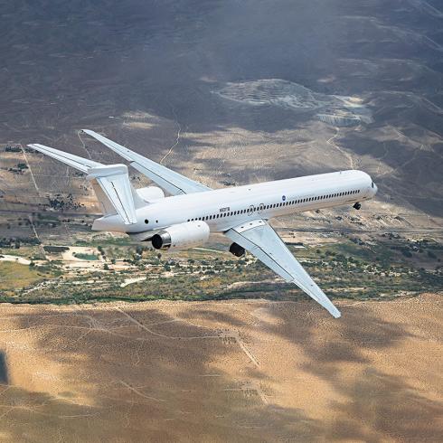 An airplane in flight. The airliner is white and has very few markings. It is flying over  low mountains, the ground is mostly brown with some sparse green foliage.