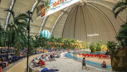 A massive brown dome covers a beachfront resort, with green tropical trees and sunbathers.