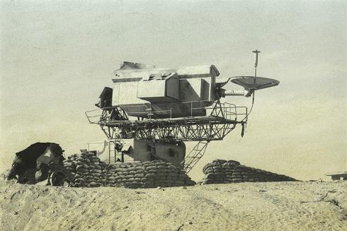 A black and white photo shows a rectangular device with a small dish in the desert.