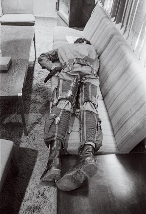 The pilot in this black-and-white photo is wearing his flight suit and sleeping face down on a couch.