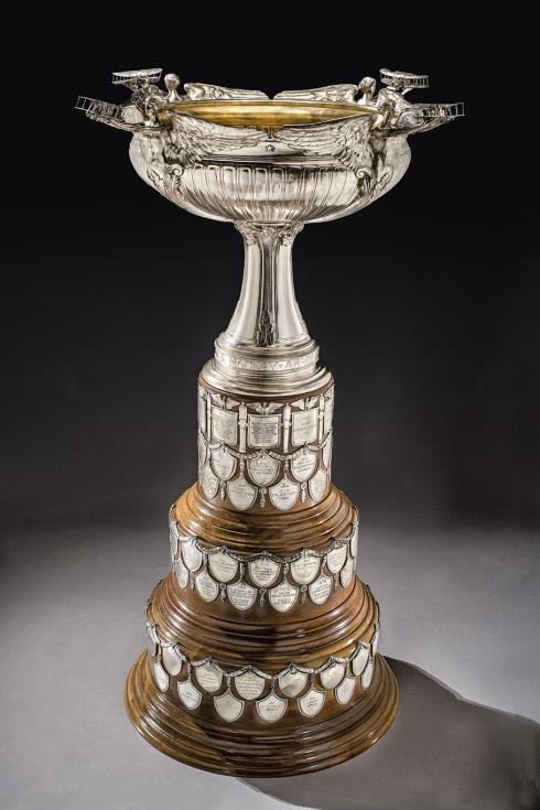 An ornate sterling silver trophy sits atop an expanding base made from three layers of polished mahogany wood. Multiple engraved metal shields are affixed to the mahogany layers. An ornate cup sits above the adorned base. 