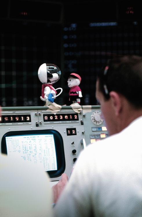 Small Snoopy, a cartoon dog, and Charlie Brown, a cartoon boy, dolls sit on top of a monitor at mission control. The Snoopy doll wears a spacesuit. 