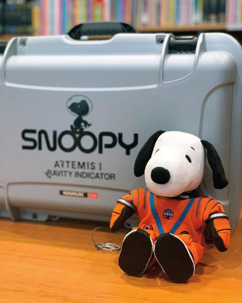 A dog-shaped Snoopy stuffed toy wears an orange flightsuit while sitting against a case labelled "Snoopy Artemis I Gravity Indicator" with a silohette of Snoopy on it. 