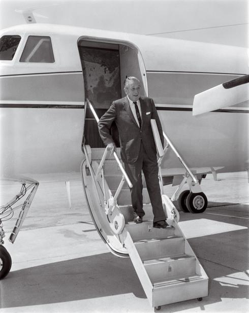 A senior man in a suit, Walt Disney, descends the staircase of his Gulfstream aircraft.