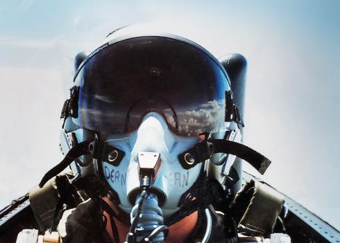 A close-up shot of a pilot sitting in the cockpit of a military jet covered by a clear plexiglas canopy. The pilot is wearing a white helmet with a dark visor and a breathing hose. White clouds are reflected by the visor.