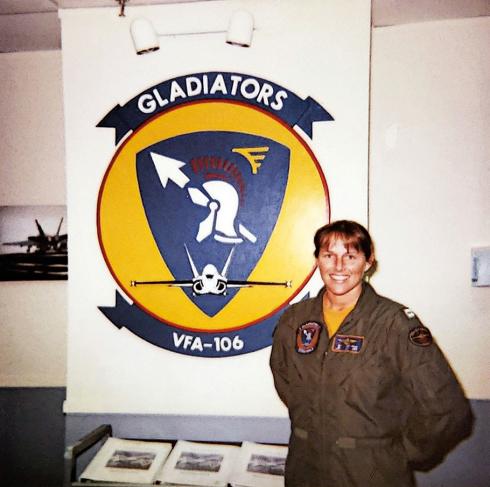 A young woman wearing an olive-green flightsuit stands next to a sign on the wall that represents U.S. Navy fighter squadron VFA-106.