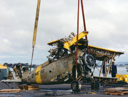 A wrecked biplane, barely recognizable and covered with seaweed, is lowered to the ground with a crane,