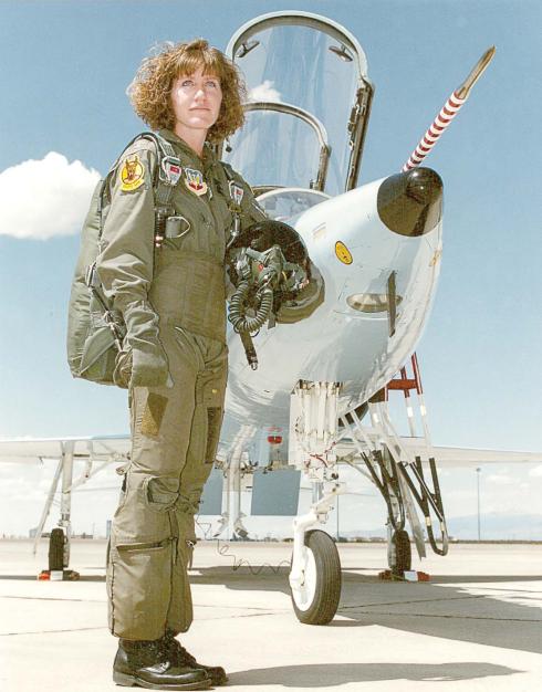 A woman in her mid-20s, wearing a pilot jumpsuit, stands in front of a military aircraft.