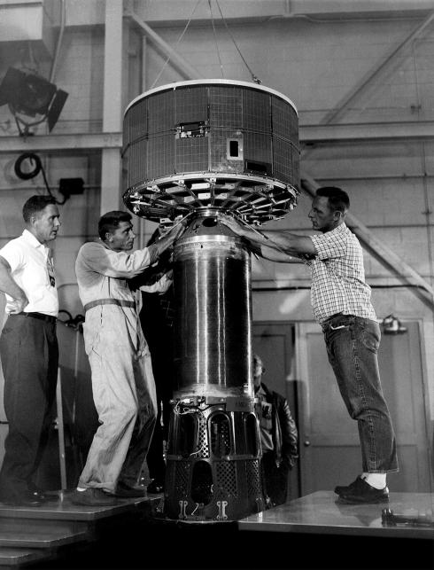 In this black-and-white image, three white men make adjustments to a large, metallic satellite inside a hangar. The satellite has a cylindrical base topped by a large disc-shape. The men are dressed casually in button-down shirts and jeans and khakis.