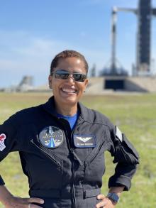 Sian Proctor stands in front of launch pad wearing astronaut jumpsuit with mission patches on it