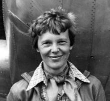 A black-and-white photo of Amelia Earhart, smiling for the camera, wearing a flight jacket. Her hair is cut short and tousled.