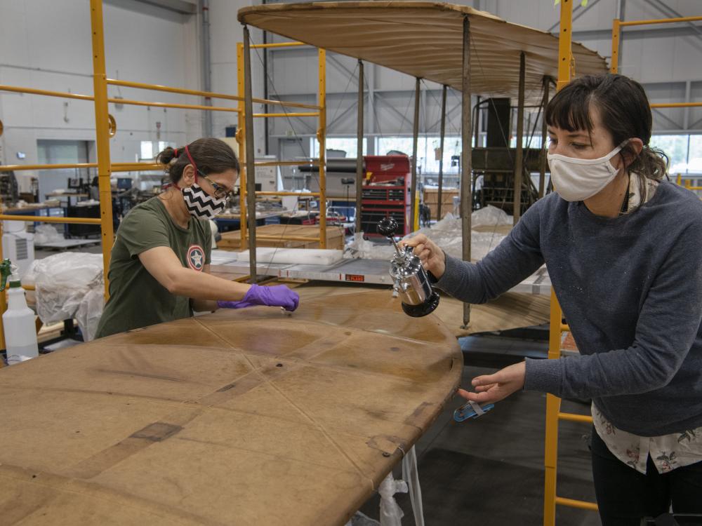 Two conservators, wearing masks, work on the Wright Flyer. 