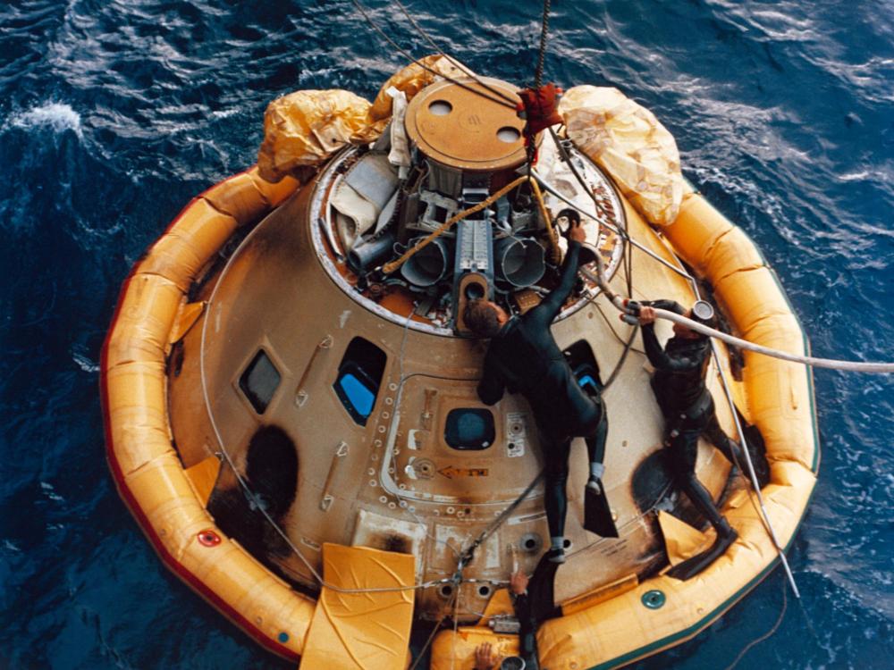 A color photograph of navy divers recovering the Apollo 6 Command Module from the Pacific Ocean. The Command Module floats on the surface of the water.