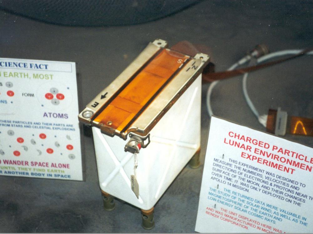 Photograph of the charged particle lunar environment experiment, which is shaped like a small rectangular box that is mostly white with orange on top. Two placards are next to the experiment to explain its function. 