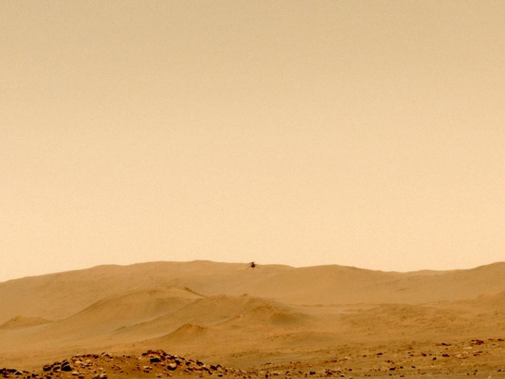 A view of the orange surface of Mars with hills in the background and a barely visible device flying above in the air.