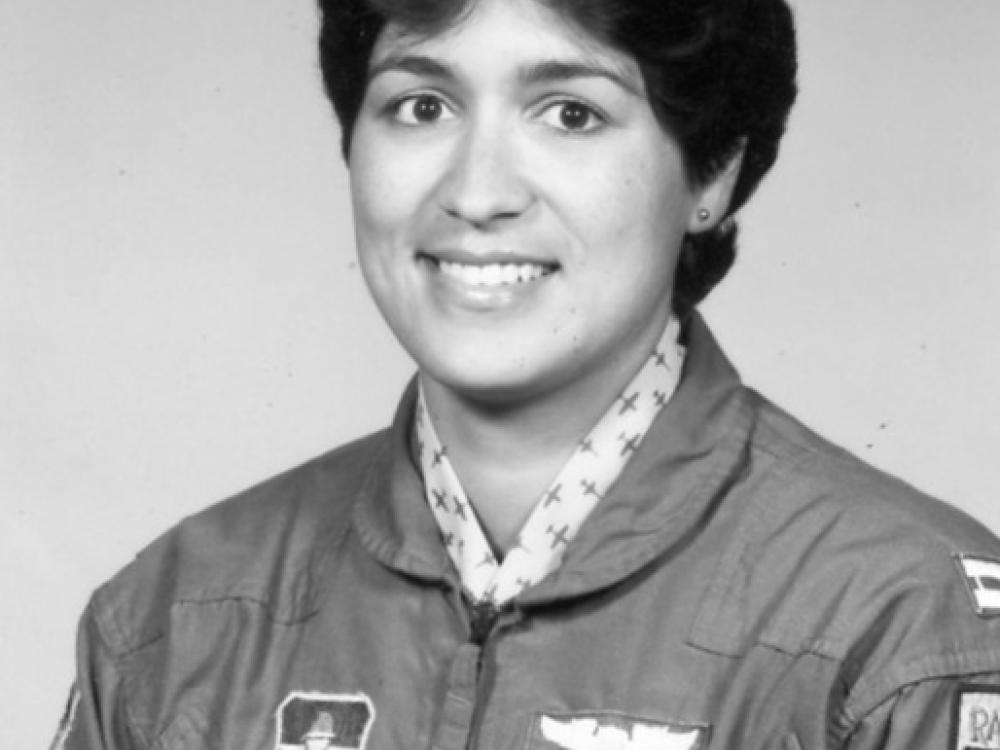 Black and white portrait of woman with short wavy hair, parted in the middle.  Stud earring in left ear (right ear is not visible).  She is wearing a dark jacket with a zipper with a cloth pull.  Patch on her right side (the left of the image) features a icon with wings.  Two patches on her left side (right of photo). Top is wings. Bottom is a nameplate that says "Custodio."  There are two pens in her left sleeve. Another patch is cut off by the edge. White collar with a dark airplane print under the jacket