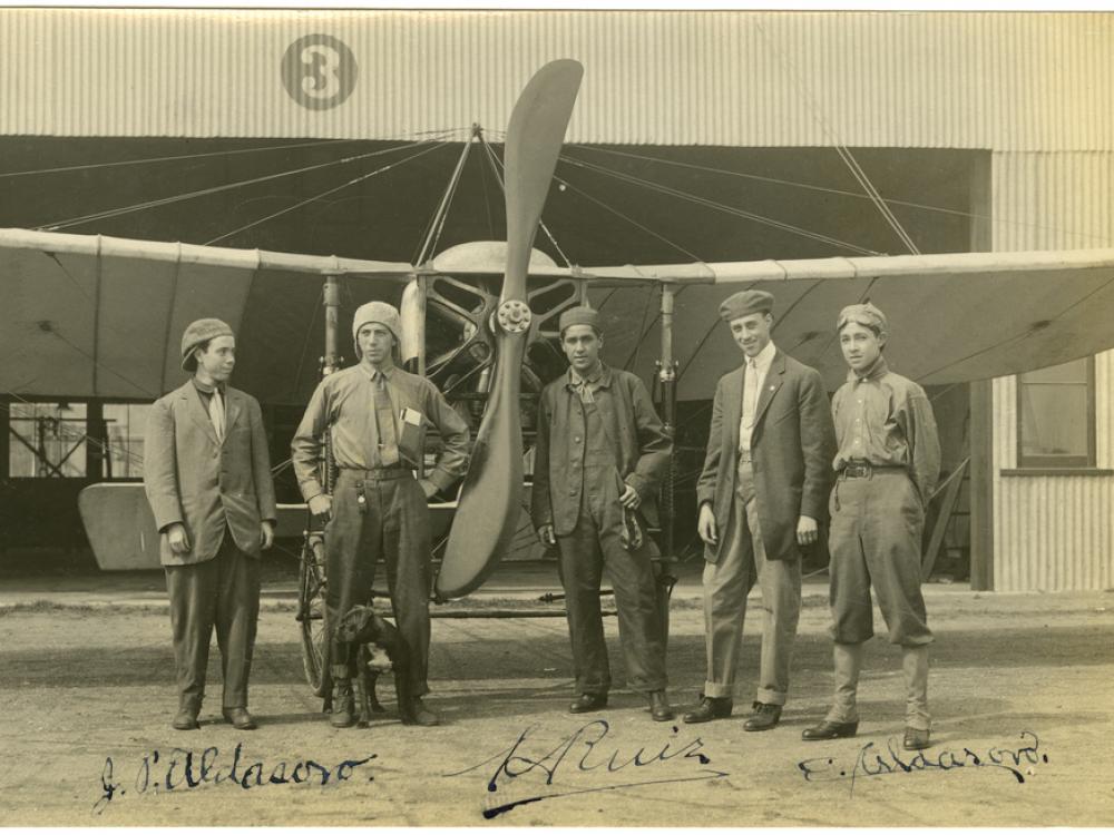 Five men stand in front of a monoplane with a two blade propeller. Background: a hanger made of corrugated metal and to the left of center is the number 3 inside a circle.  From left to right: first man in a suit jacket facing slightly to the right with a backwards cap. second man is wearing a backwards cap and does not wear a jacket and has his left hand in his pocket. The third man is on the other side of the propeller is wearing a cap and unbuttoned jacket.  The fifth man stands with his hands behind him
