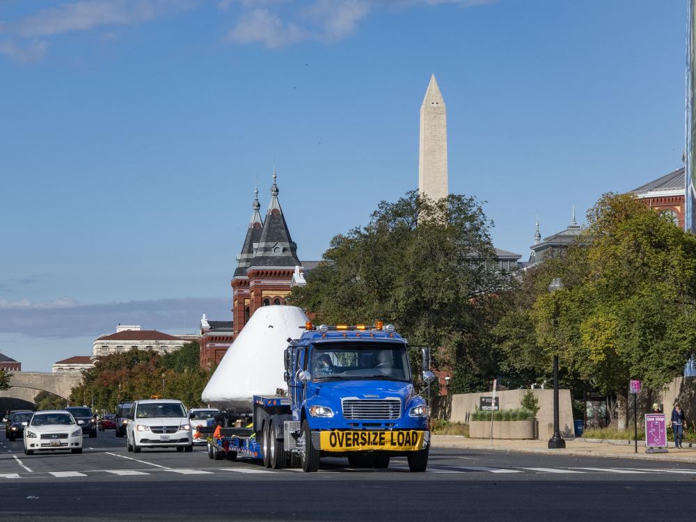 A truck on a public road with other vehicles behind it as carries a large white triangular object with the city view in the background.