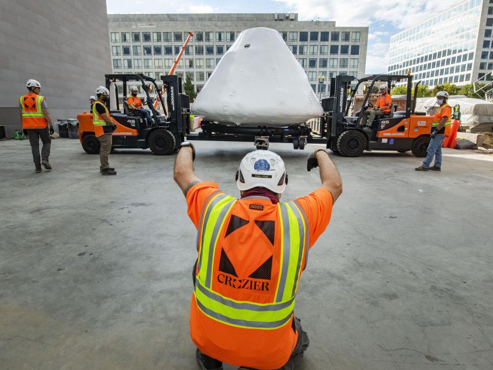 Apollo 11 command module Columbia being lifted by two forklifts and surrounded by workers.