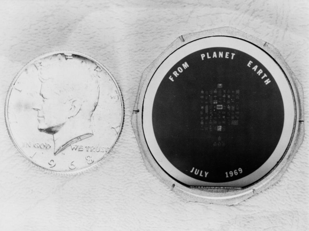 A black and white photograph of a circular disc with "from planet Earth, July 1969" written in large font. Smaller type is in the middle of the disc, but is not legible. The disc is next to a quarter for scale, and is roughly the same size.