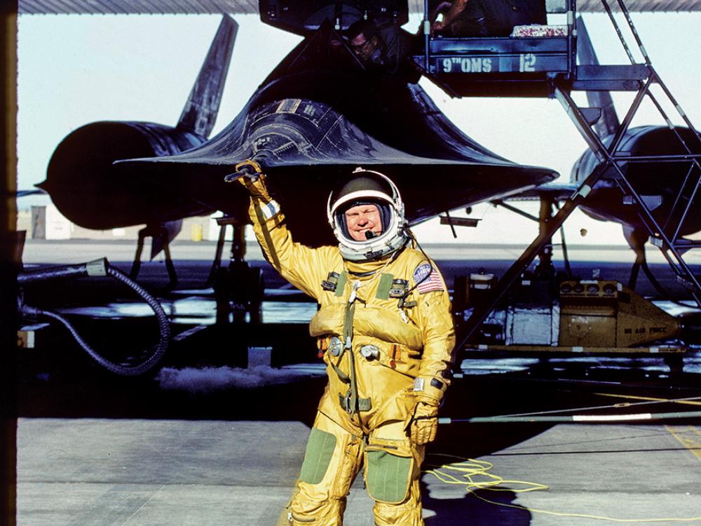 A man in a yellow suit and a space helmet points to the Blackbird airplane behind him. 