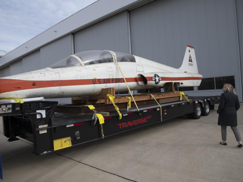 A woman examines a T-38 Talon aircraft while it's strapped to the back of a truck without its landing gears.