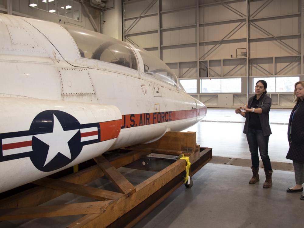 Two women examine a T-38 Talon aircraft while it's strapped to the back of a truck without its landing gears.