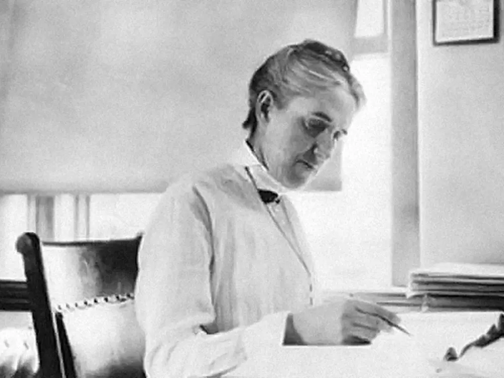Black and white image of Henrietta Swan Leavitt sitting at a desk and photographed from a side angle. She is writing with a pen in her hand.