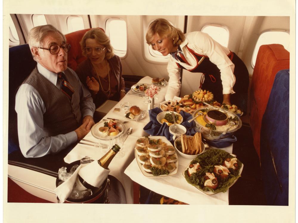 A color photo of a flight attendant serving food to two passengers aboard a plane.