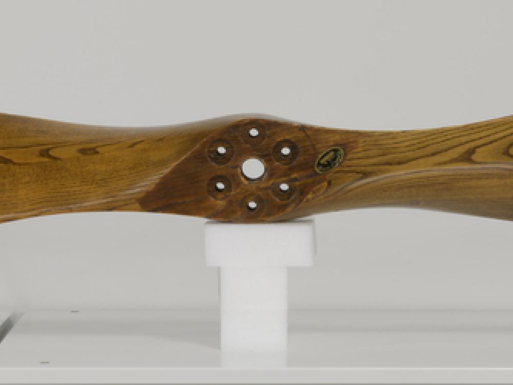 A varnished wood propeller on a white stand against an off-white background.