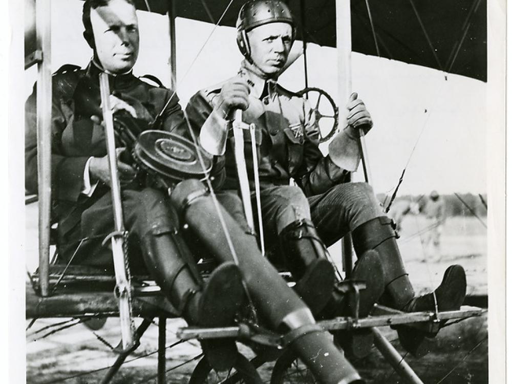 Two men in the seat of a Wright airplane. The man on the left is holding a large machine gun and the man on the right is at the controls.