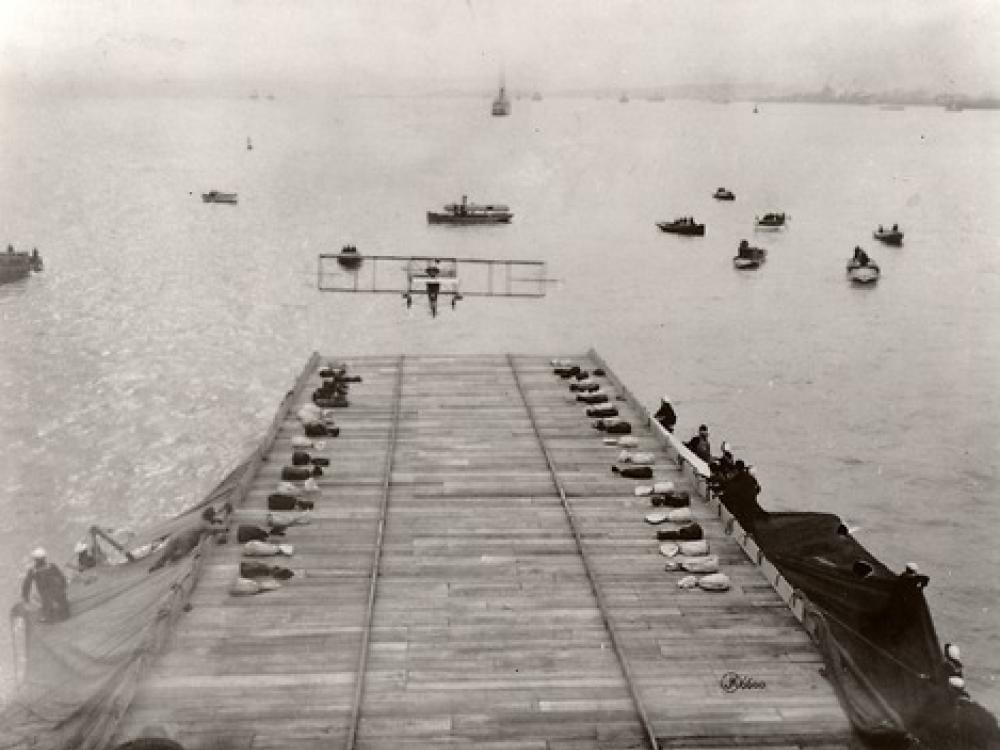 A Curtiss Model D flying toward a ship which has a makeshift runway. Other boats dot the water.