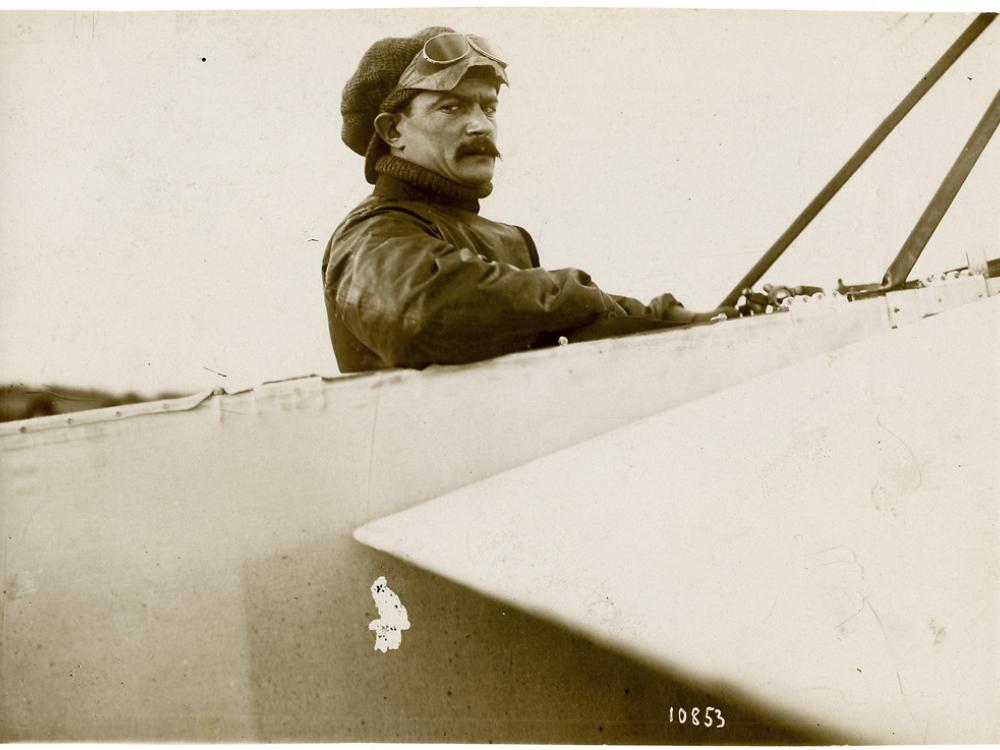 A man wearing a hat and goggles on his head, sitting in the cockpit of an airplane, looking toward the camera.