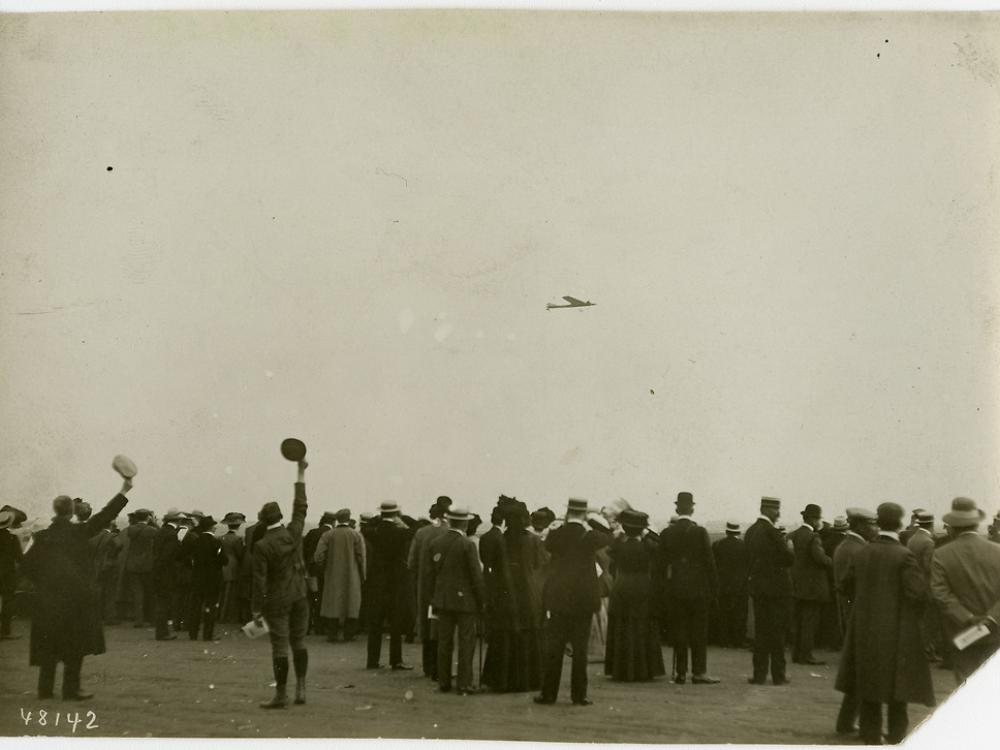 A crowd of people, some cheering, looking toward the sky where an airplane can be seen in the distance.