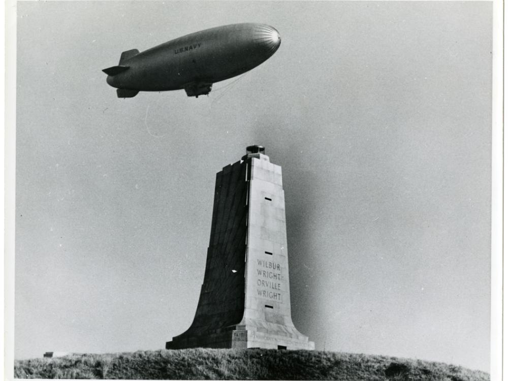 One-half right front view of a U.S. Navy airship in flight over the square column-shaped Wright brothers memorial, which has both brothers names inscribed on it.