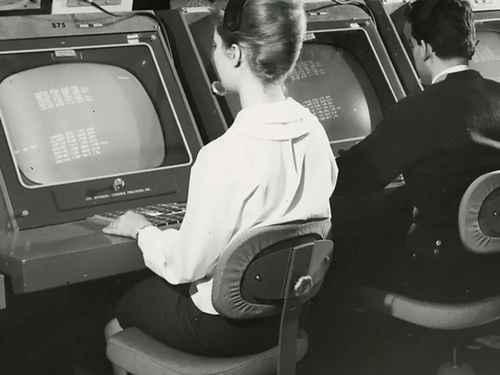Two people sit at computer terminals, facing the screen working. 