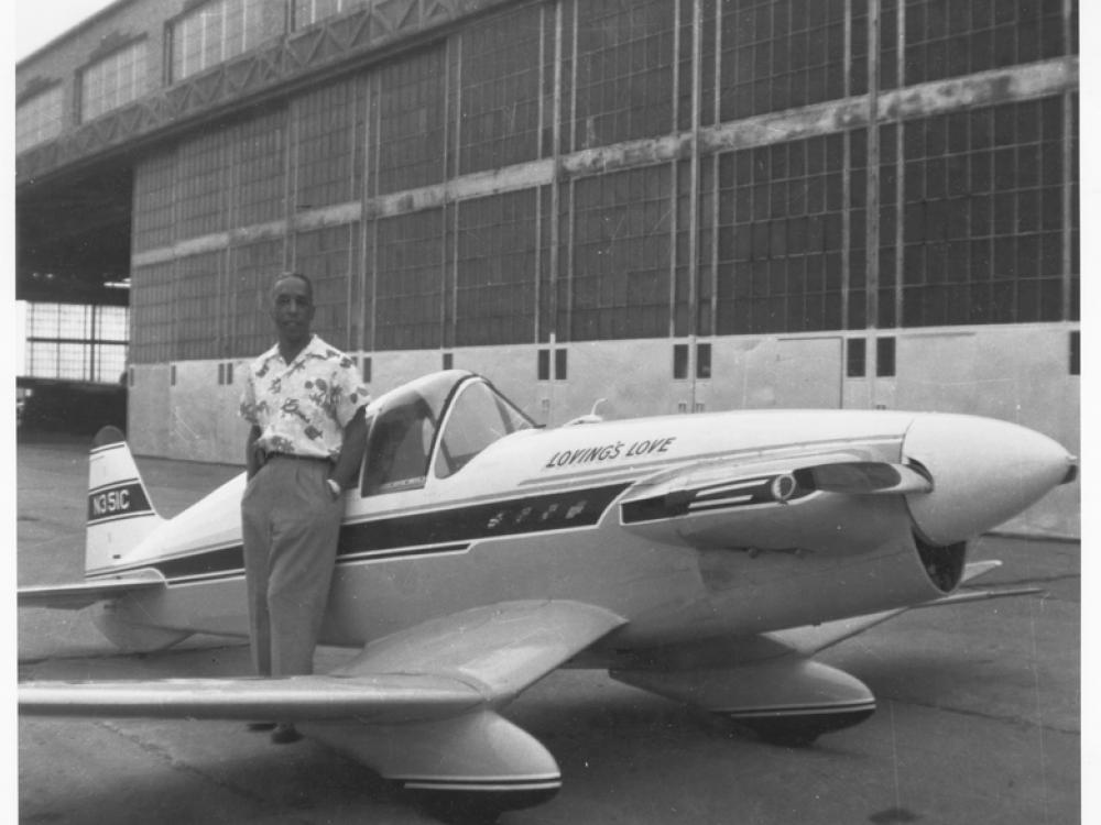 A standing person poses next to a small monoplane in front of an airplane hangar. 
