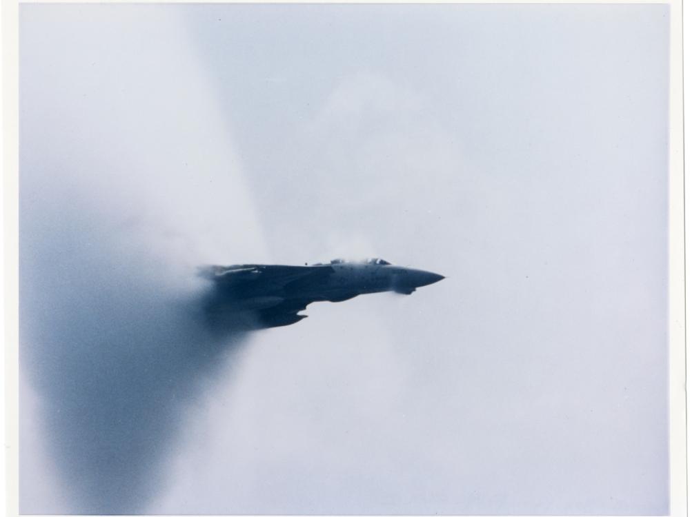 A jet aircraft emerging from a cone shaped cloud of vapor in the air. 