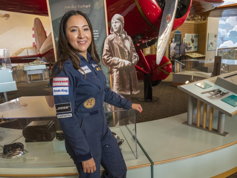 A woman in a flight uniform poses next to a bright colored early aircraft in a museum setting.